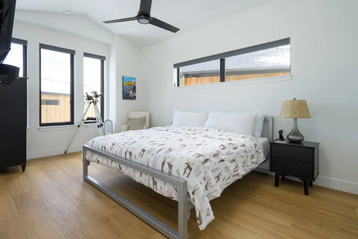 A peaceful bedroom with a plush bed featuring a patterned comforter, flanked by a nightstand and lamp. Ample windows fill the room with natural light, creating a soothing ambiance ideal for rest.