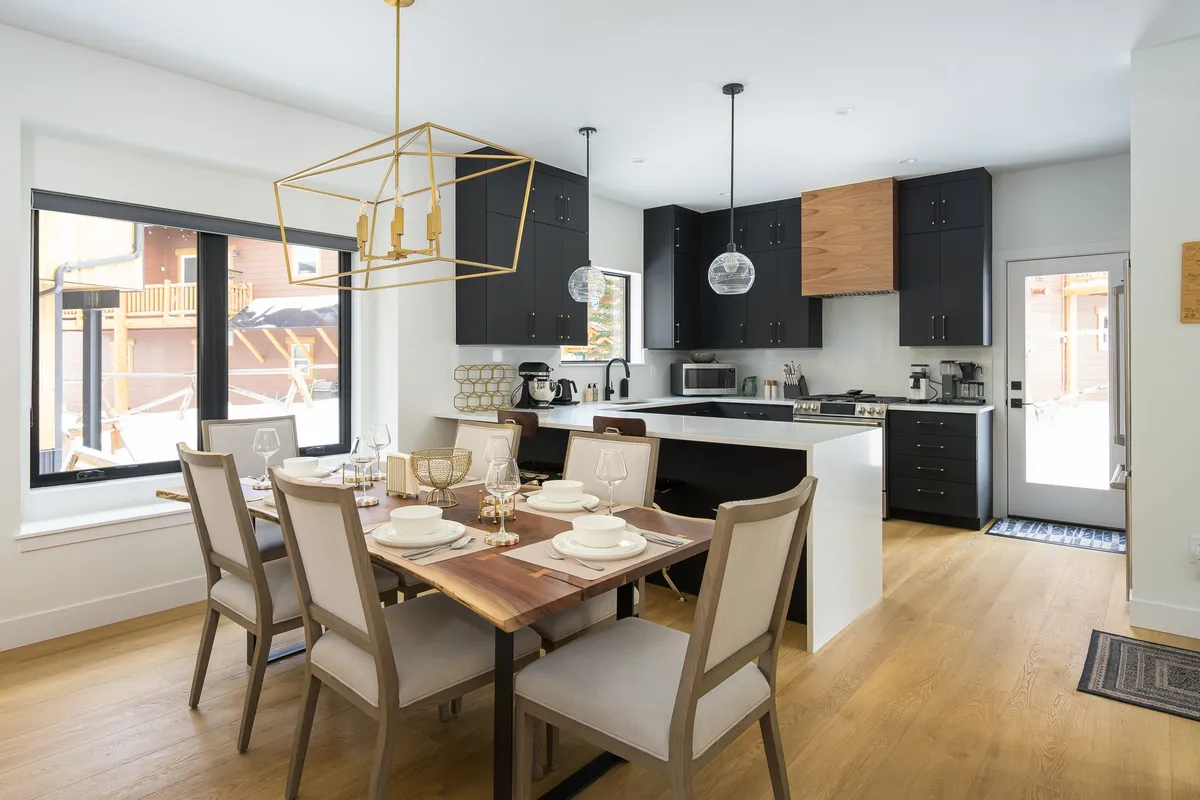 A bright and inviting open-concept dining area featuring a live-edge dining table set for six, a kitchen island, and large windows that flood the space with natural light. Hanging light fixtures add a touch of elegance and warmth, while the setup is perfect for family meals and entertaining guests.