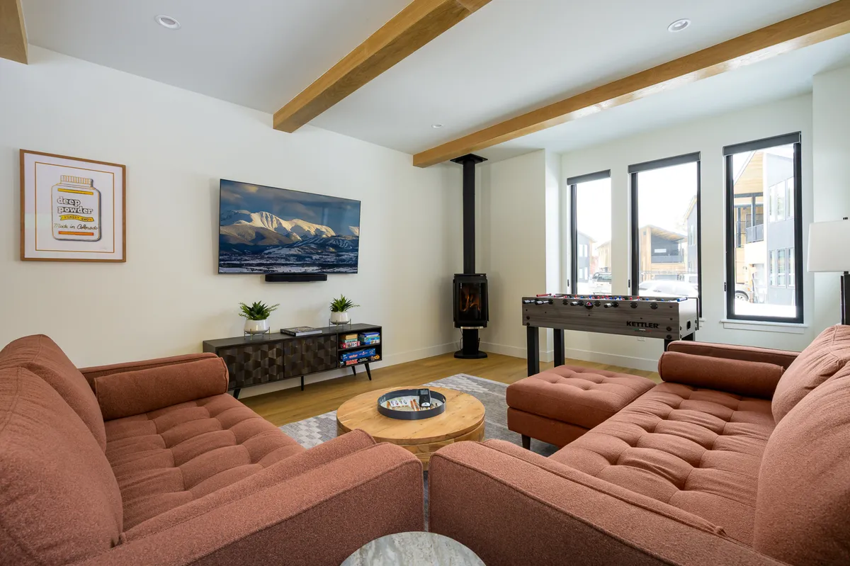 This image showcases a bright and airy living room featuring a plush sofa, a large 65" Smart TV, a striking fireplace, a foosball table, and a central coffee table. The space is designed for both relaxation and entertainment, with ample seating and natural light streaming through expansive windows.