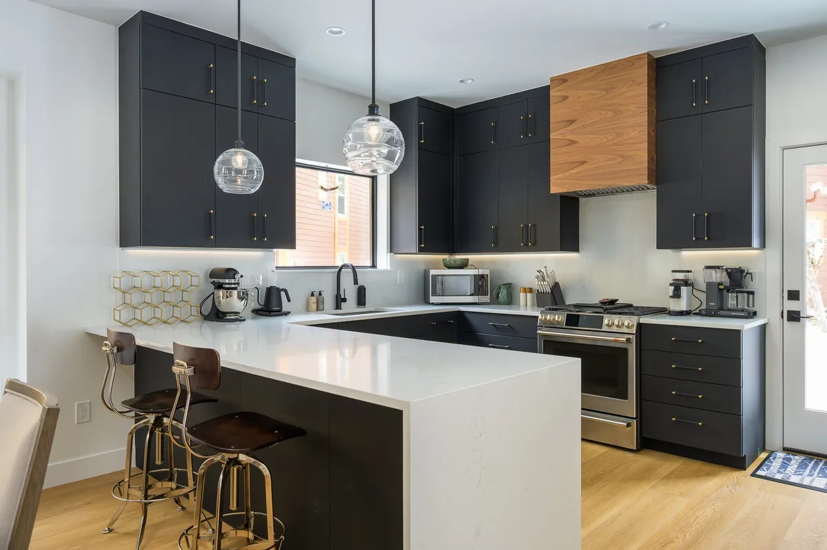 This image features a modern kitchen with a large island countertop, sleek black cabinetry, wood accents, and state-of-the-art appliances. The space is illuminated by stylish lighting fixtures, and the overall atmosphere is contemporary and functional, ideal for both cooking and entertaining.