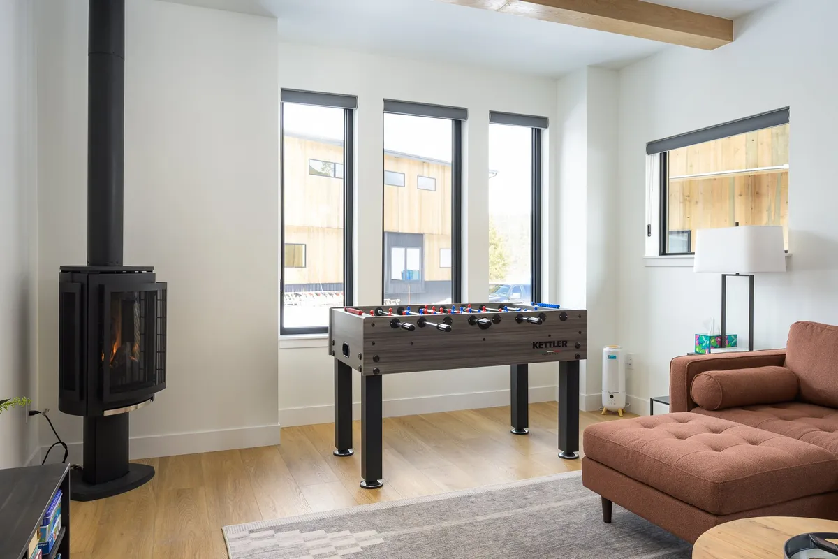 This image zeroes in on the entertainment side of the living space, highlighting a foosball table, a roaring fireplace, a comfy couch, and panoramic windows offering views of the alpine landscape. The mood is playful yet cozy, perfect for friendly matches or unwinding by the fire.