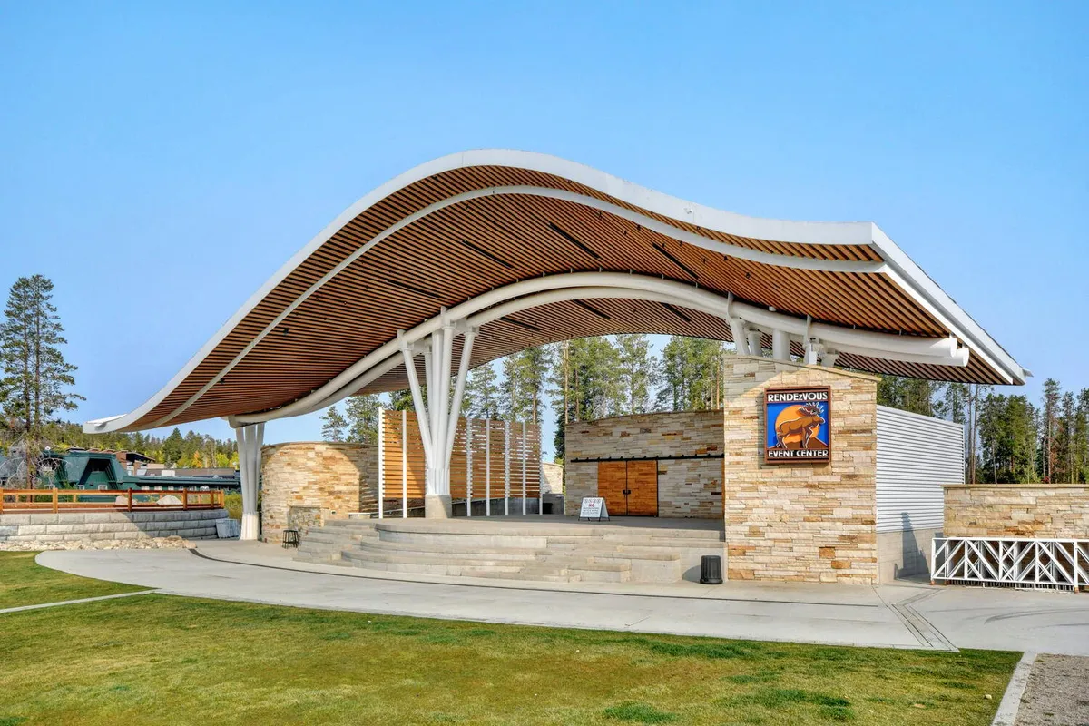 An exterior shot of a unique curved roof structure with stone walls, open stage area, and a backdrop of trees and blue skies. The scene is set for outdoor gatherings, concerts, and community events, suggesting lively neighborhood activities and the spirit of local adventure.