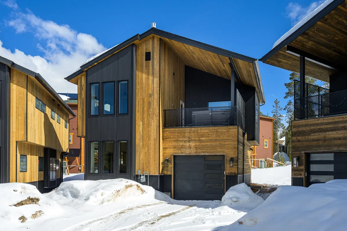 The image showcases a contemporary single-family villa with striking modern architecture, featuring a blend of wood and metal exteriors and expansive windows. The setting highlights a spacious balcony perfect for watching mountain sunsets, and a cozy fire pit area ideal for gathering after a day of nearby snow adventures. Snow blankets the surrounding landscape, evoking a serene and inviting winter escape.